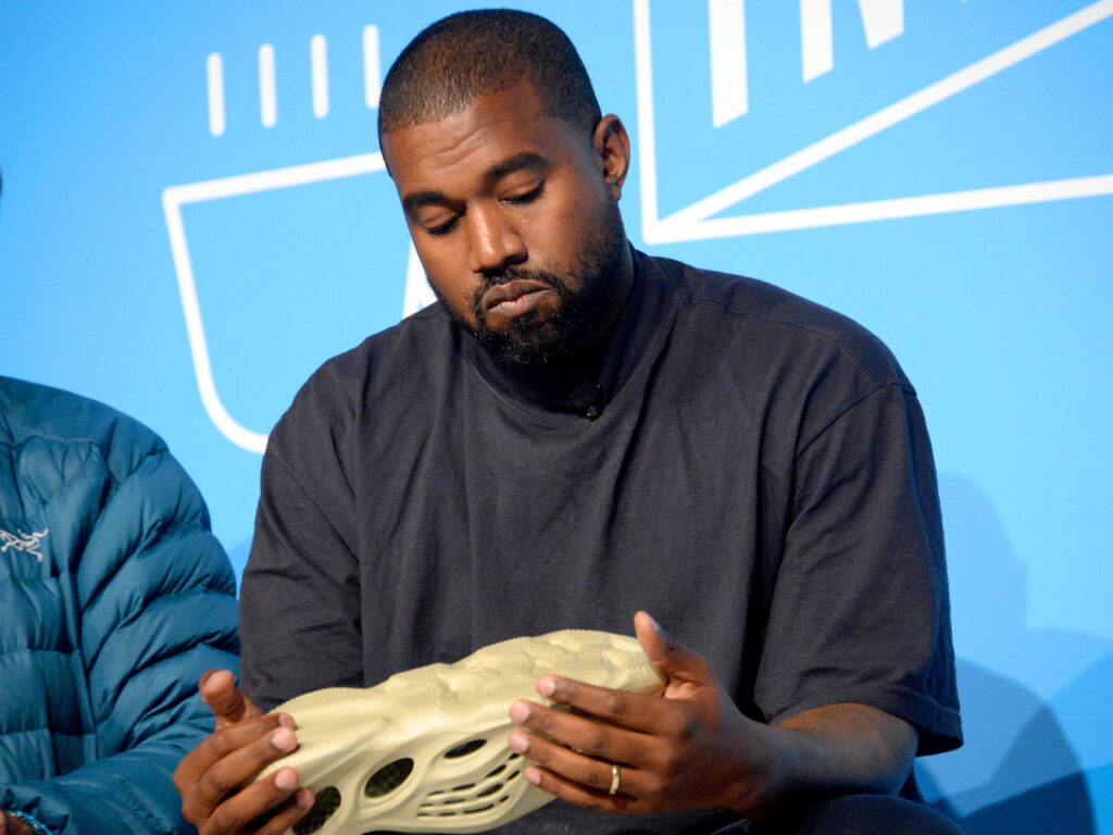 Kanye West holds a cream-colored sneaker against a blue backdrop