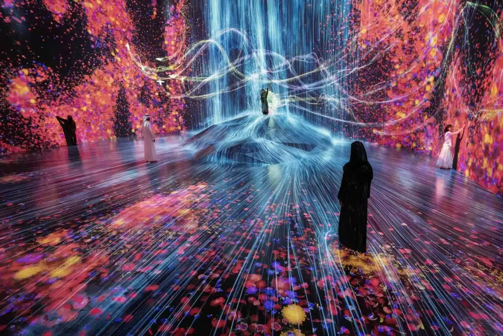 Visitors walk through an immersive digital art installation of cascading blue light and colorful flowers surrounding a central mound