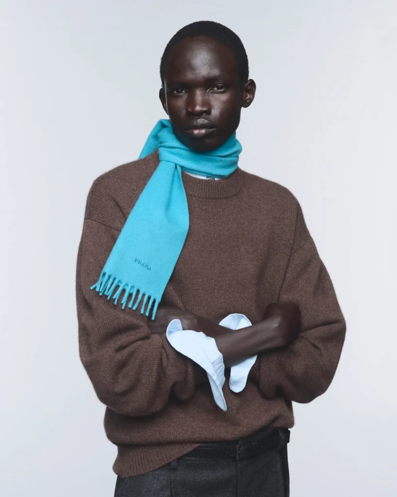 Model wearing a brown Re-Cashmere sweater and turquoise Prada scarf, holding a white garment against a light background