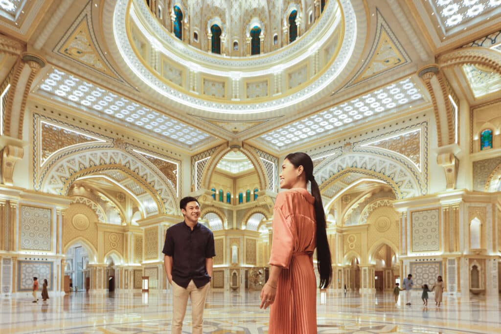 Two people admire the ornate, brightly lit interior of Qasr Al Watan with intricate arches and stained-glass windows