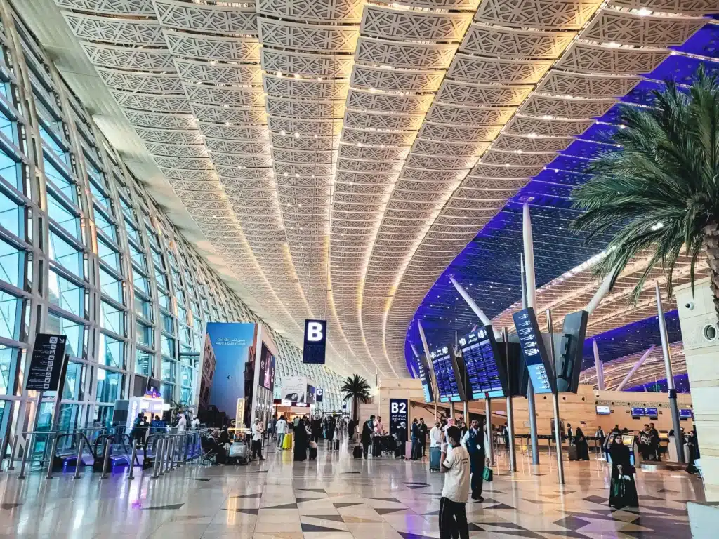 Spacious airport terminal with patterned ceilings, palm trees, flight information screens, and travelers with luggage