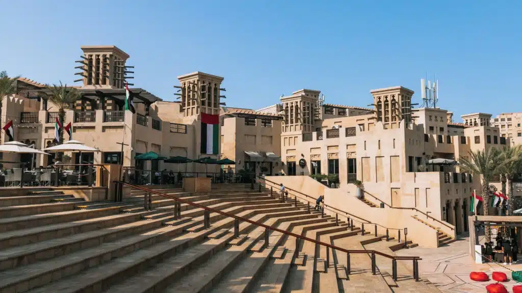 Traditional Middle Eastern-style market plaza with wind towers, broad steps, palm trees, outdoor seating, and UAE flags