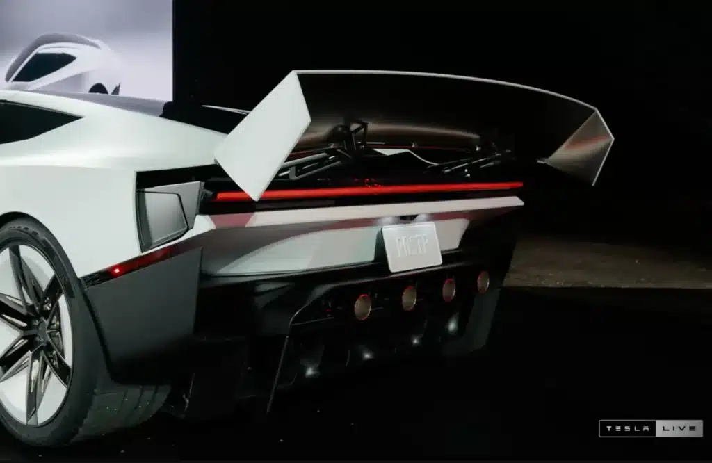 Rear view of a futuristic white Tesla vehicle with an open cargo area, red taillight, and large aerodynamic wing