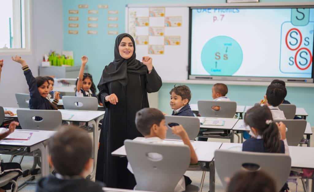 A teacher leads a phonics lesson while children raise their hands in a classroom
