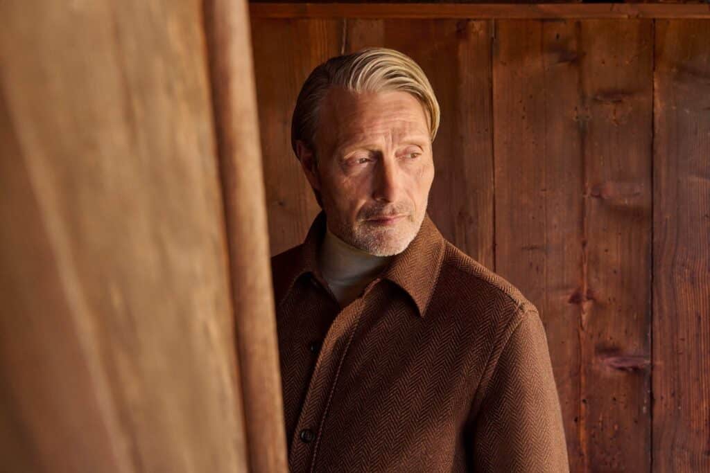 Actor Mads Mikkelsen in a Zegna brown coat looks through a doorway against a wooden wall
