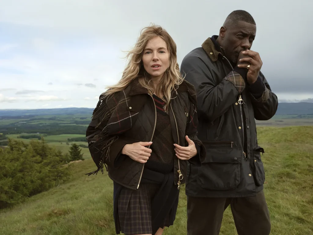 Two people, one of them is Idris Elba, stand on a grassy hillside overlooking a broad rural landscape; the man raises his hand to his mouth, and both are wearing Barbour jackets.