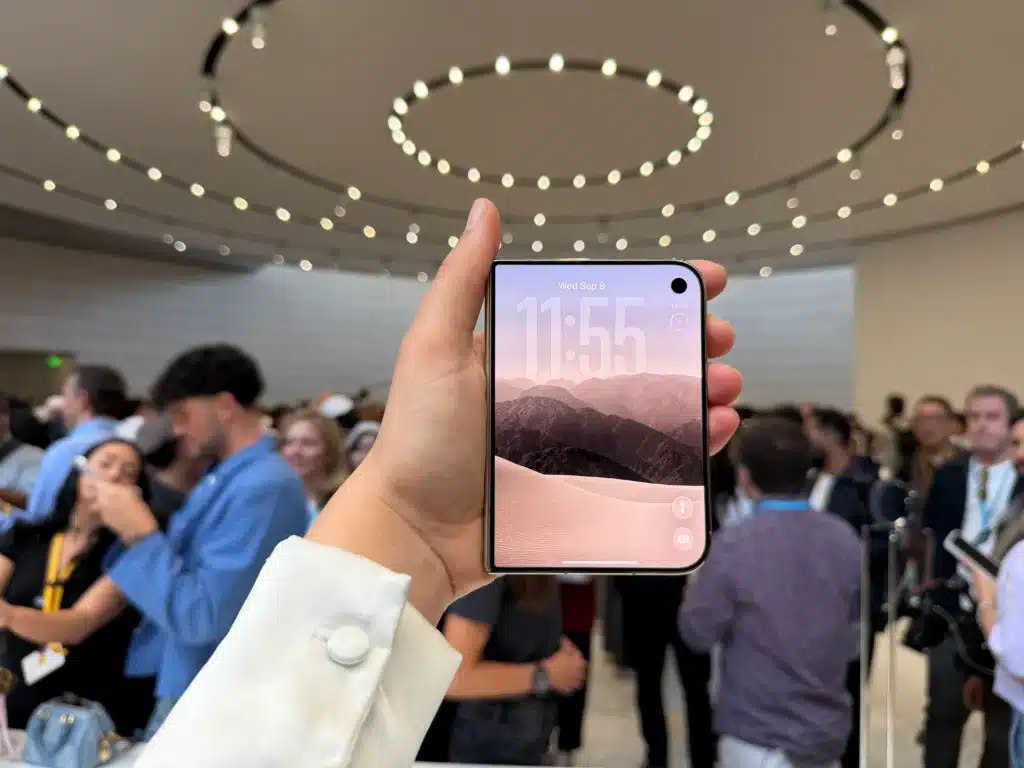 A hand holds a smartphone showing 11:55 beneath circular lights in a crowded room